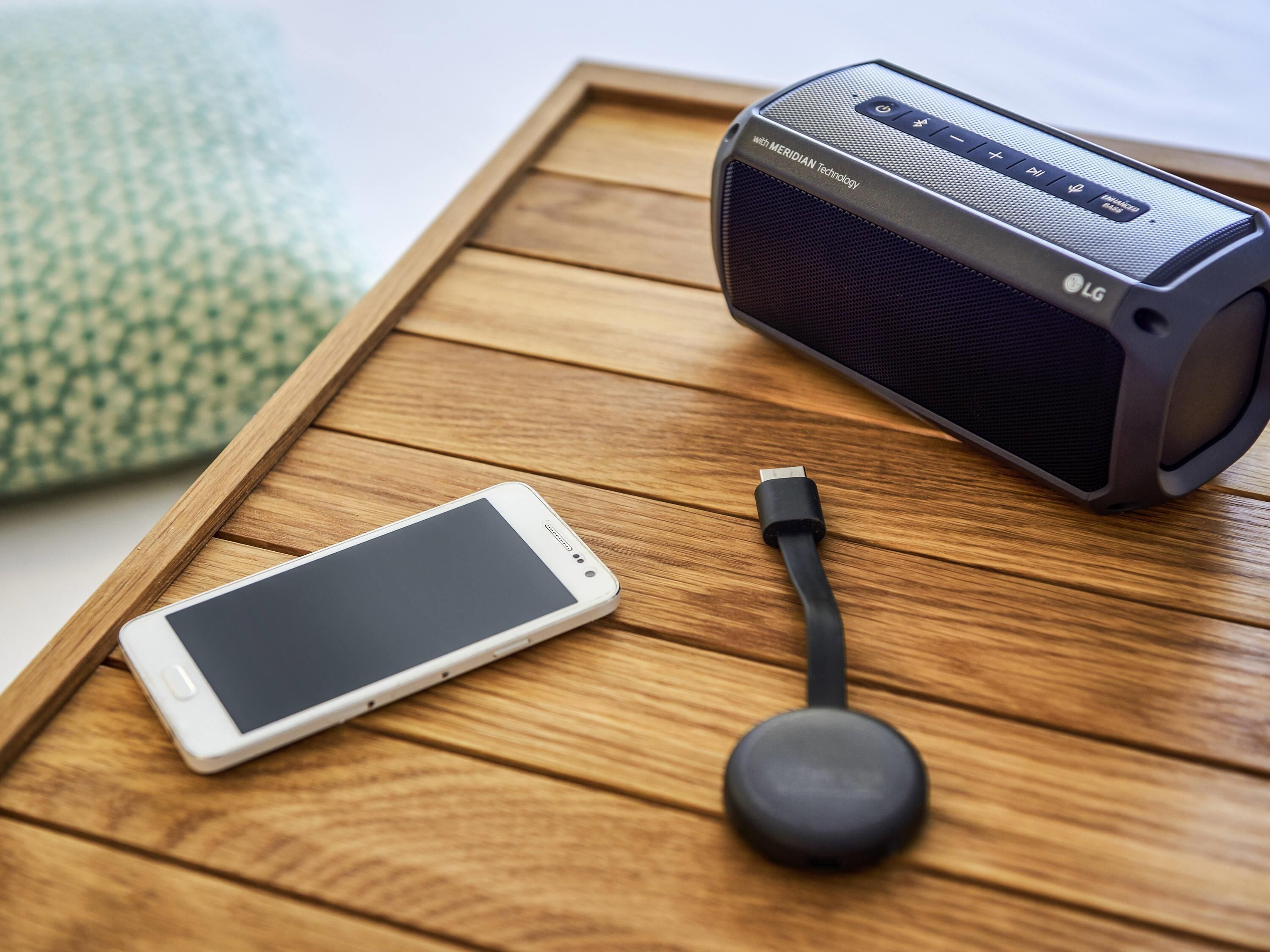 A smartphone, a black speaker, and a charging cable on a wooden tray.
