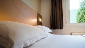 A hotel room with a neatly made bed, a headboard with integrated lighting, a window with curtains, and a view of greenery outside. - Brit Hotel Nantes St Herblain - Le Kerann (Saint-Herblain)