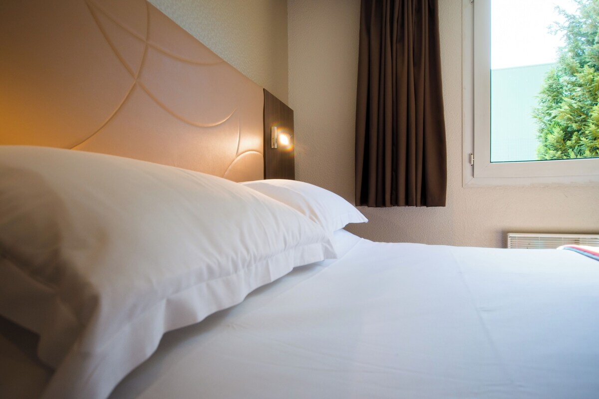 A hotel room with a neatly made bed, a headboard with integrated lighting, a window with curtains, and a view of greenery outside.