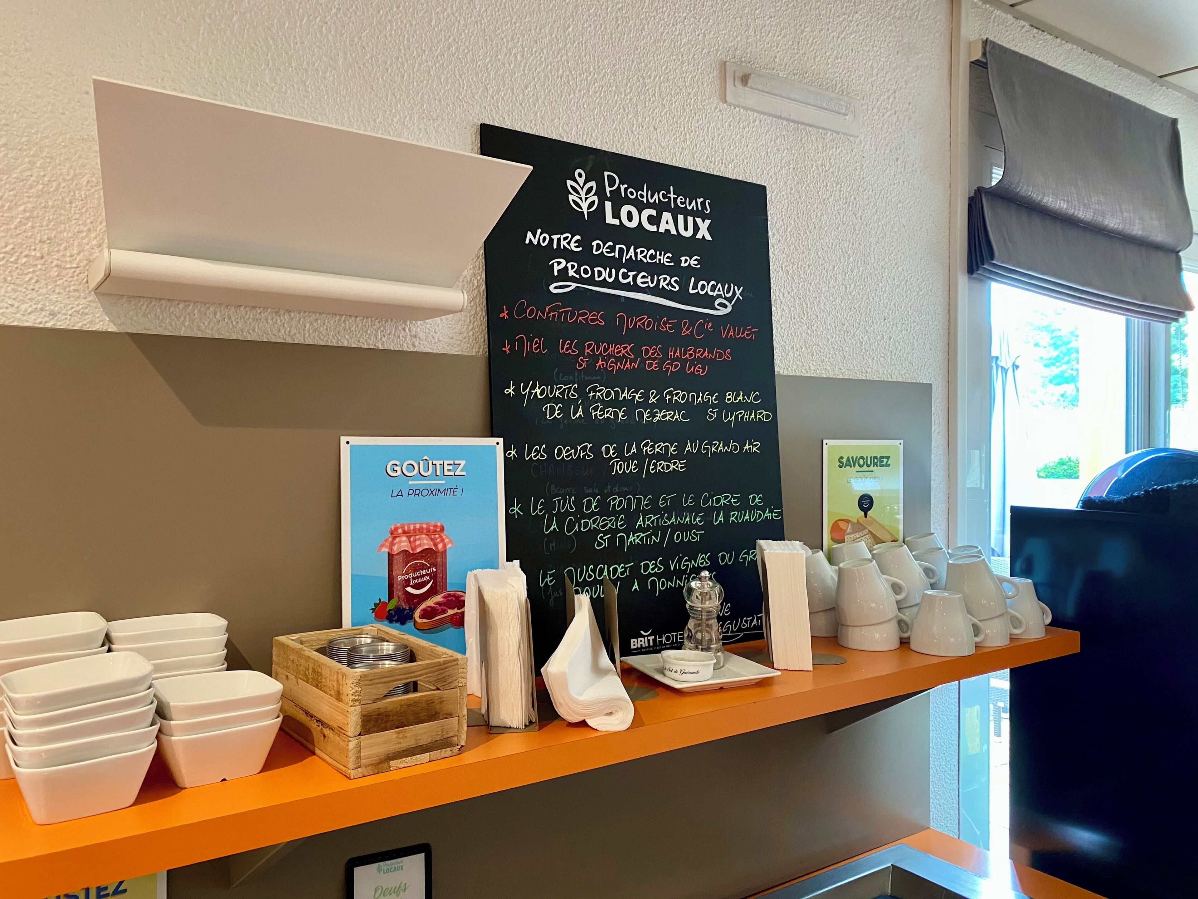 Zona de recepción de un hotel con un menú en una pizarra que anuncia productos locales, una exposición de cuencos de cerámica blanca y una ventana con vistas.