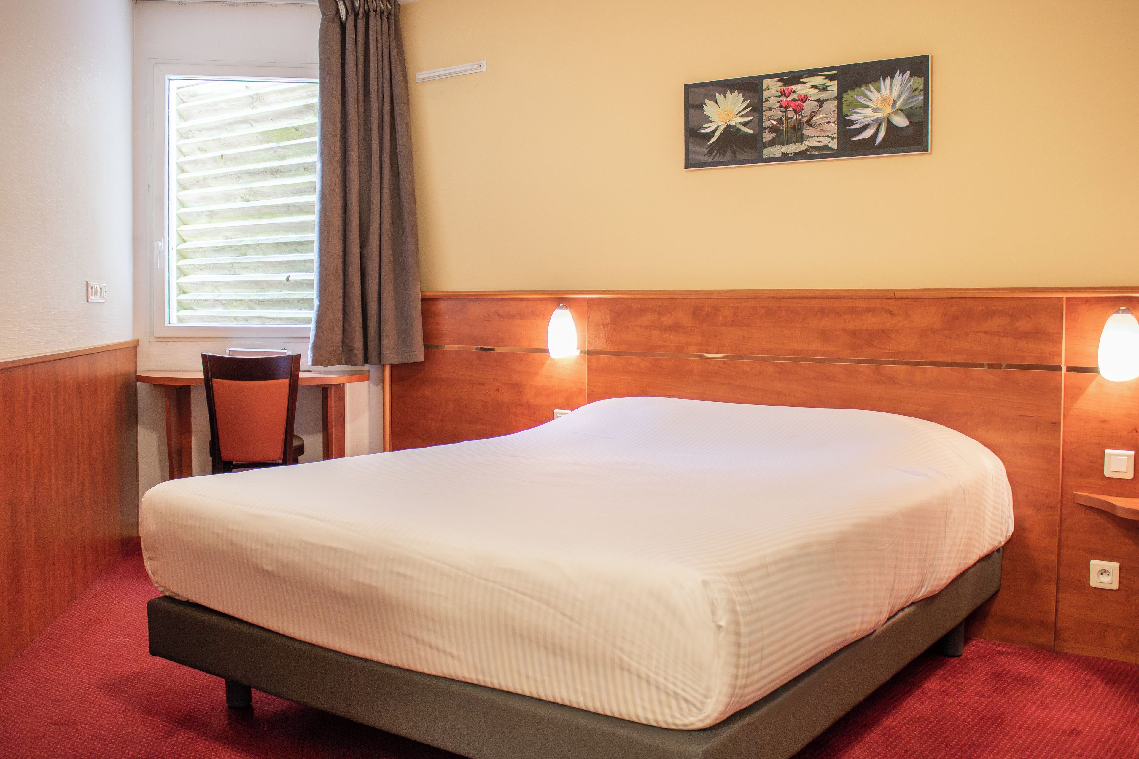 Une chambre d’hôtel comprenant un lit, un bureau, une chaise, une fenêtre avec des stores, un rideau et des lampes fixées au mur.