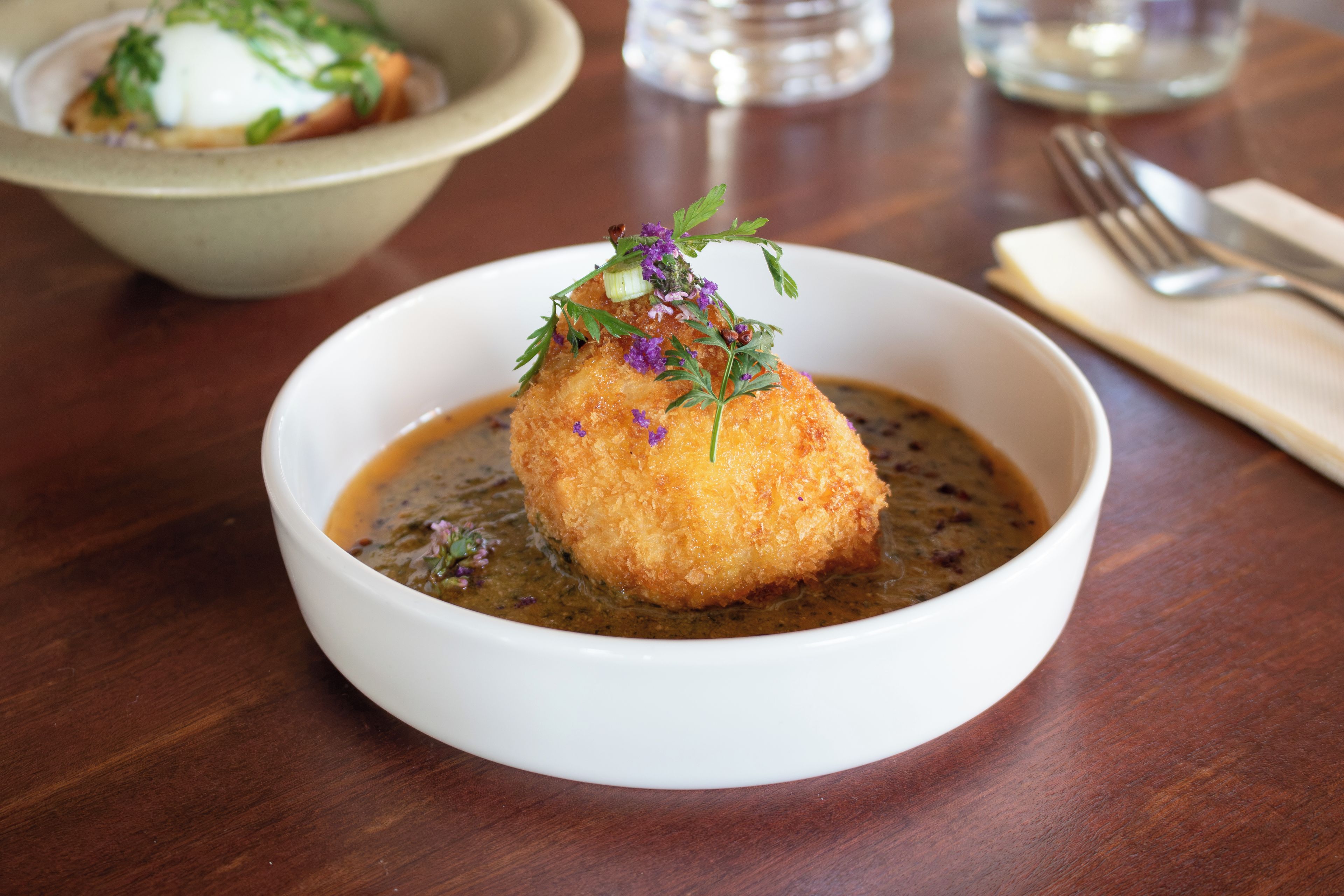 A white bowl with a single breaded item on a dark sauce, placed on a wooden table.
