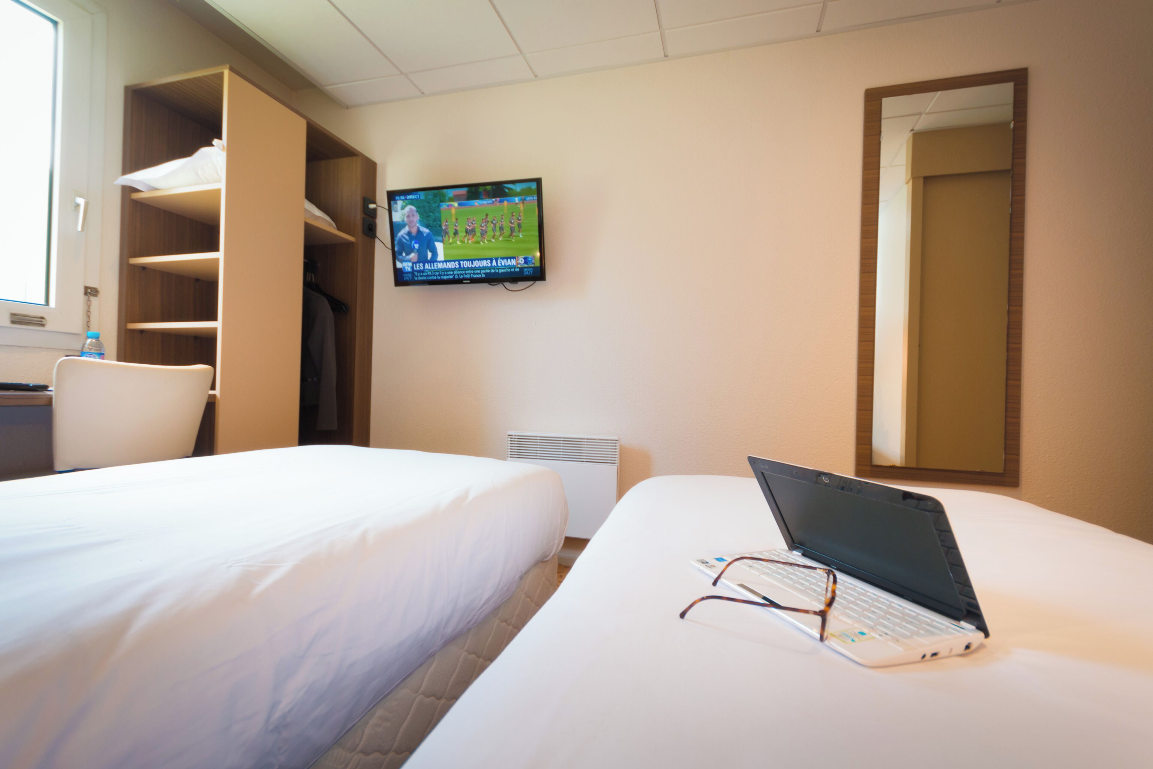 Une chambre d’hôtel avec deux lits, un téléviseur fixé au mur, un ordinateur portable posé sur l’un des lits et un miroir au mur.