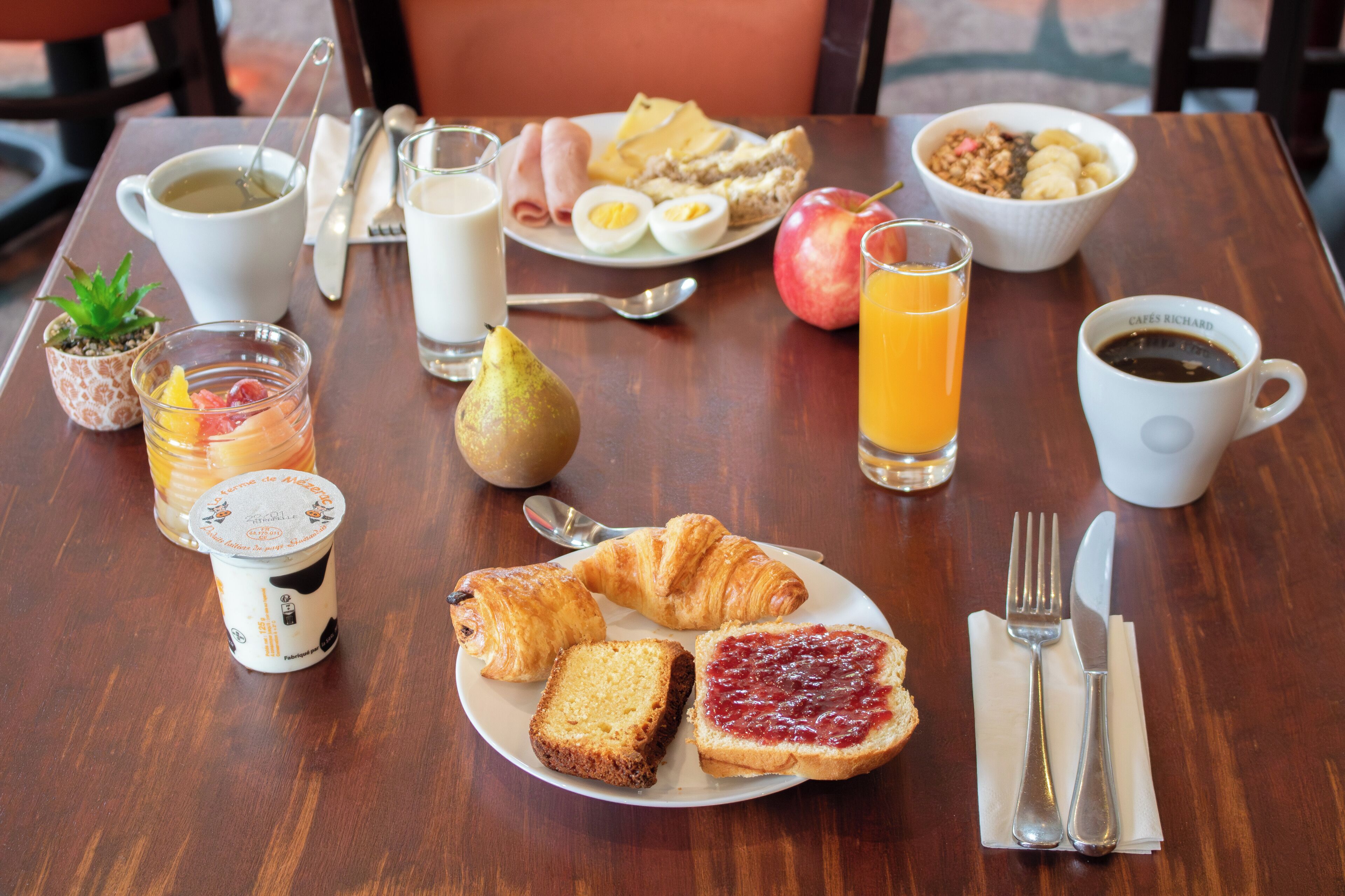 A breakfast table with croissants, toast, jam, eggs, a pear, a glass of orange juice, a cup of coffee, and a bowl of cereal.