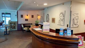 A hotel reception area with a wooden reception desk, a seating area with tables and chairs, and framed artwork on the walls. - Brit Hotel Nantes St Herblain - Le Kerann (Saint-Herblain)