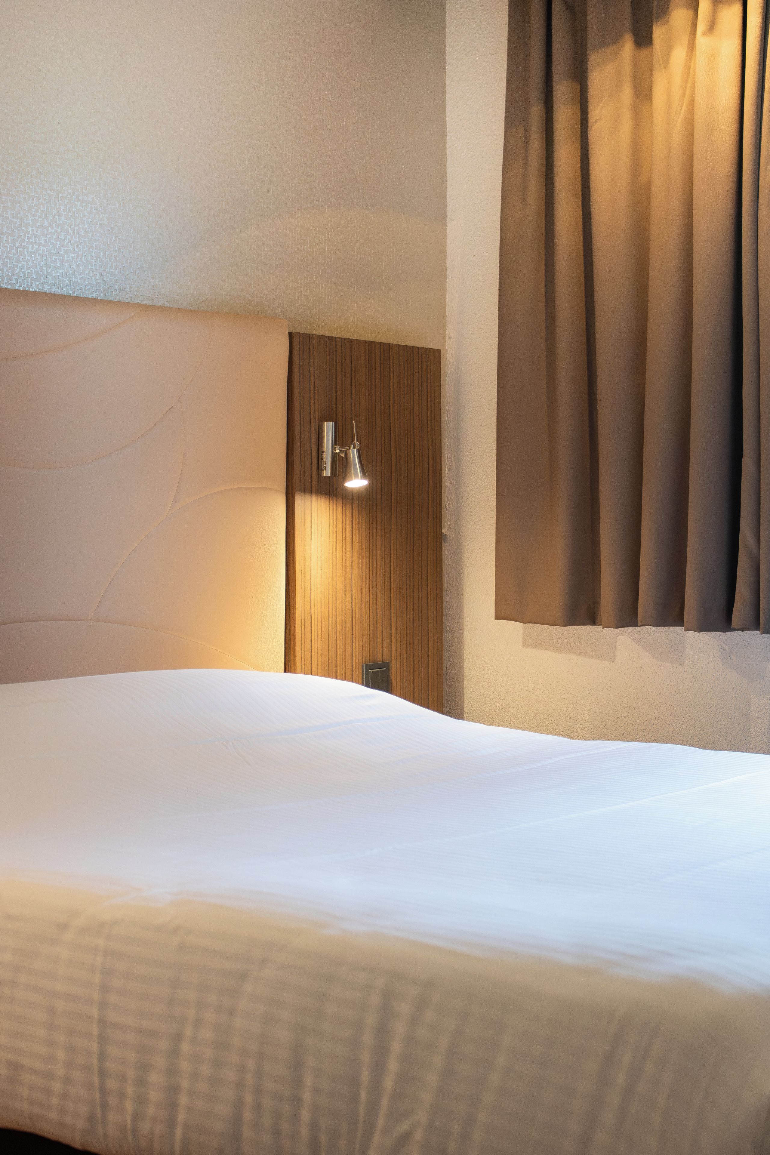 Una habitación de hotel con una cama grande, cabecera de madera, lámpara de pared y una ventana con cortinas.