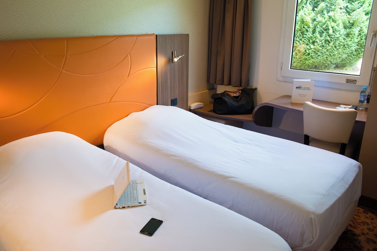 A hotel room with two beds, a desk, a chair, a phone, a laptop, and a window with a view of greenery.