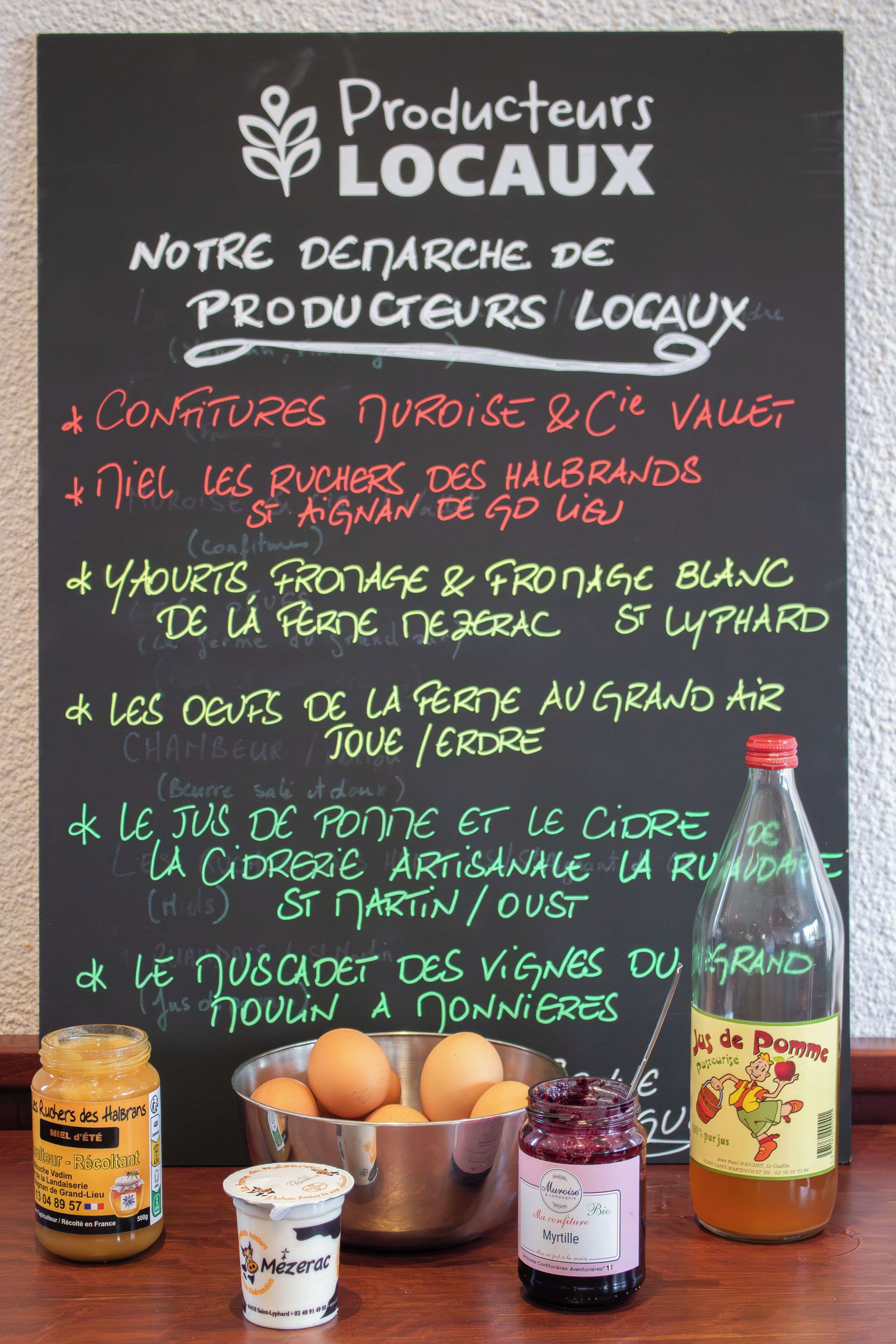 Una pizarra muestra un menú con productos locales e ingredientes alimenticios, un tarro de miel, un cuenco de huevos, una botella de mermelada y un recipiente de yogur.