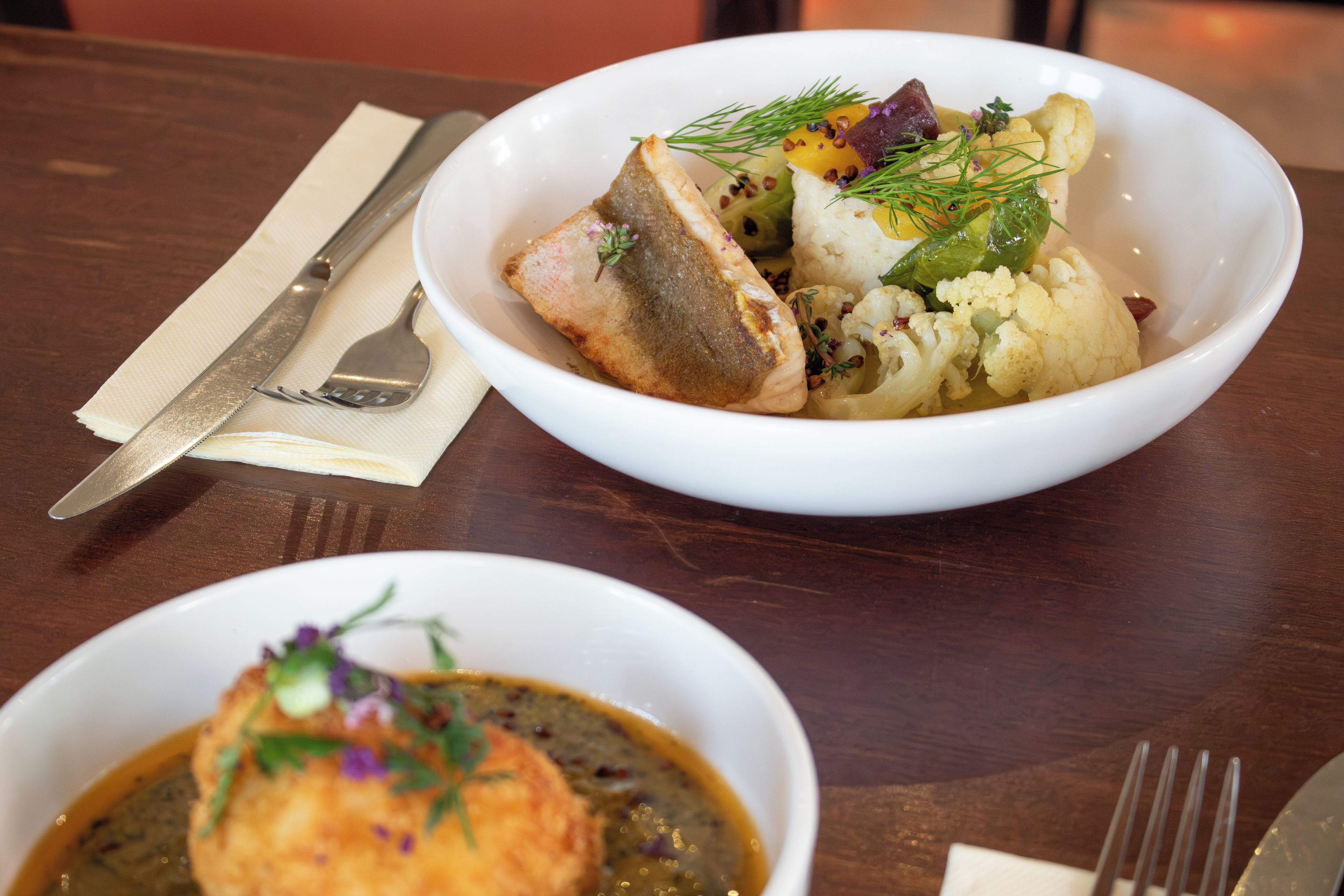 Une table à manger avec un bol blanc contenant un filet de poisson, du chou-fleur et des accompagnements, ainsi qu’un plus petit bol contenant une sauce et du poisson.