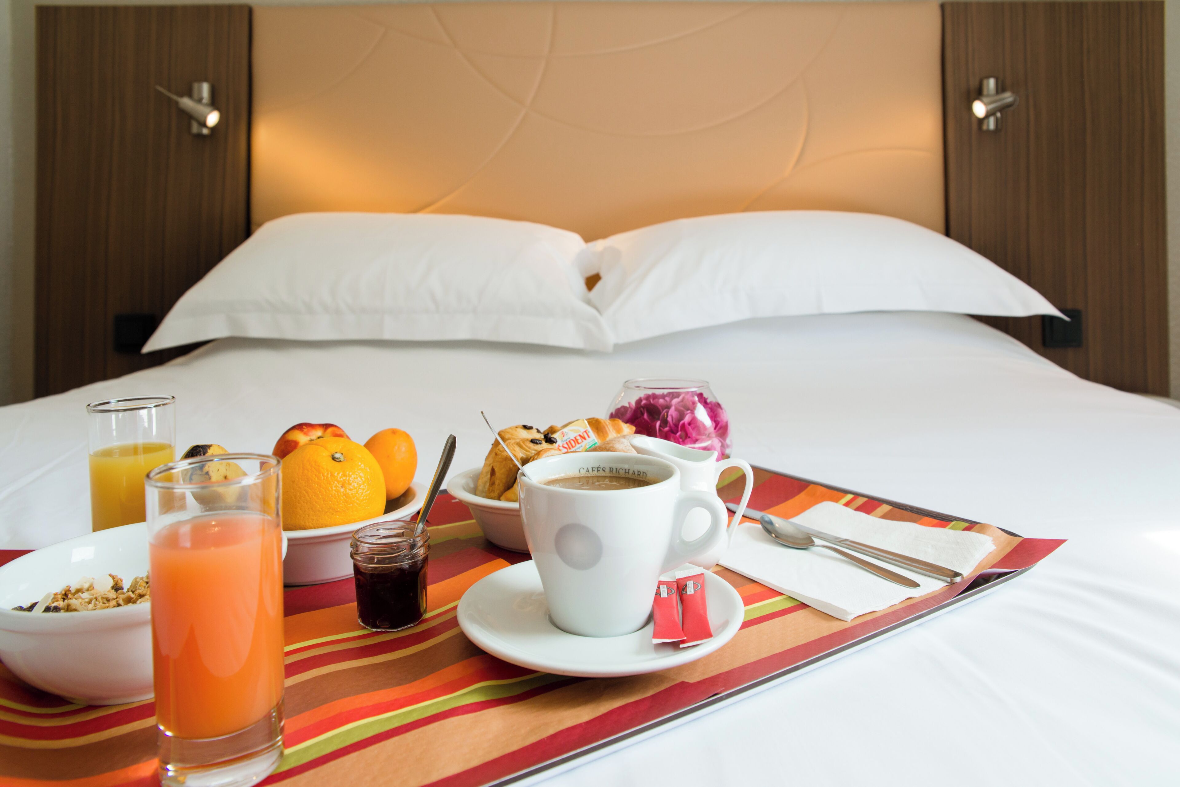 Een keurig opgemaakte ontbijtsetting op een hotelbed, met een gestreept tafelkleed, een kom met ontbijtgranen, een glas sinaasappelsap, een kop koffie en een bord gebakjes.
