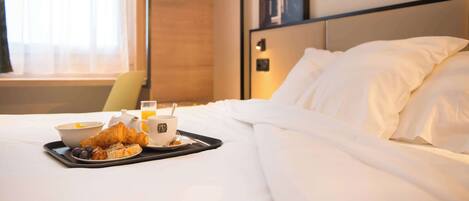 A neatly made bed with white linens, a tray with croissants, a bowl of fruit, and a cup of coffee on a hotel room table.