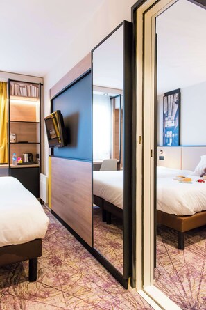 Habitación de hotel con dos camas, un televisor montado en la pared, una estantería y un espejo grande.