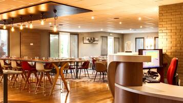 Een moderne hotellobby met een receptiebalie, meerdere eettafels met stoelen en een groot raam dat natuurlijk licht binnenlaat.