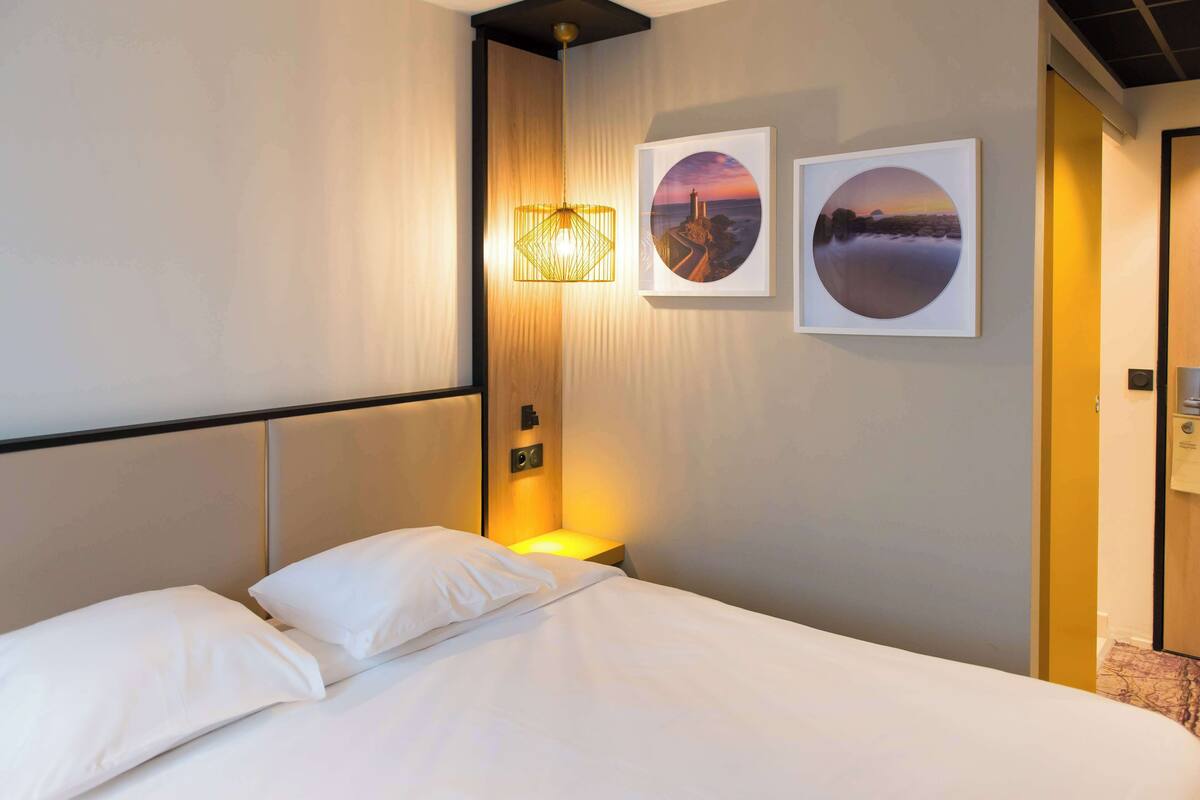 A hotel room with a neatly made bed, a wall-mounted lamp, and two framed pictures depicting coastal scenes.