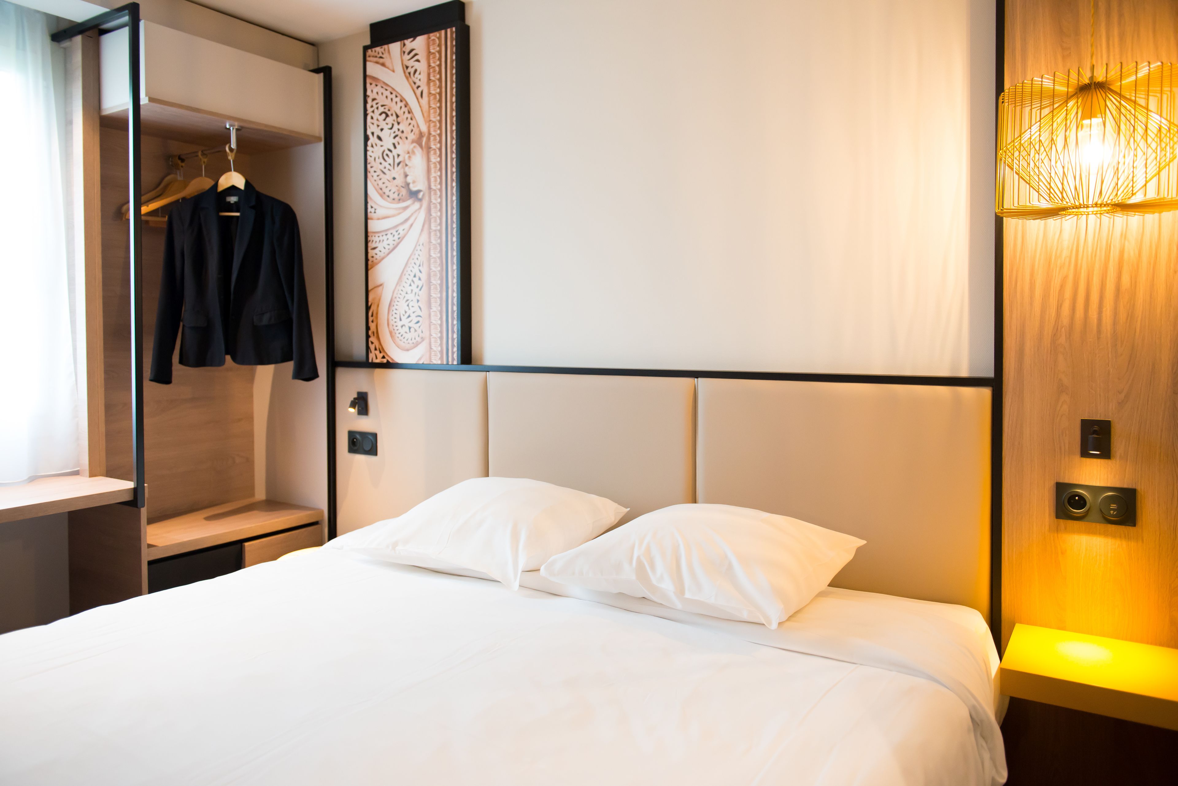 Habitación de hotel con cama, almohadas blancas, perchero y mesita de noche, además de una lámpara fijada a la pared.