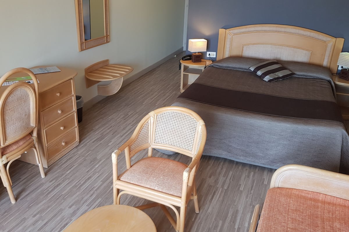 A hotel room with a bed, two chairs, a desk, a nightstand with a lamp, and a mirror.