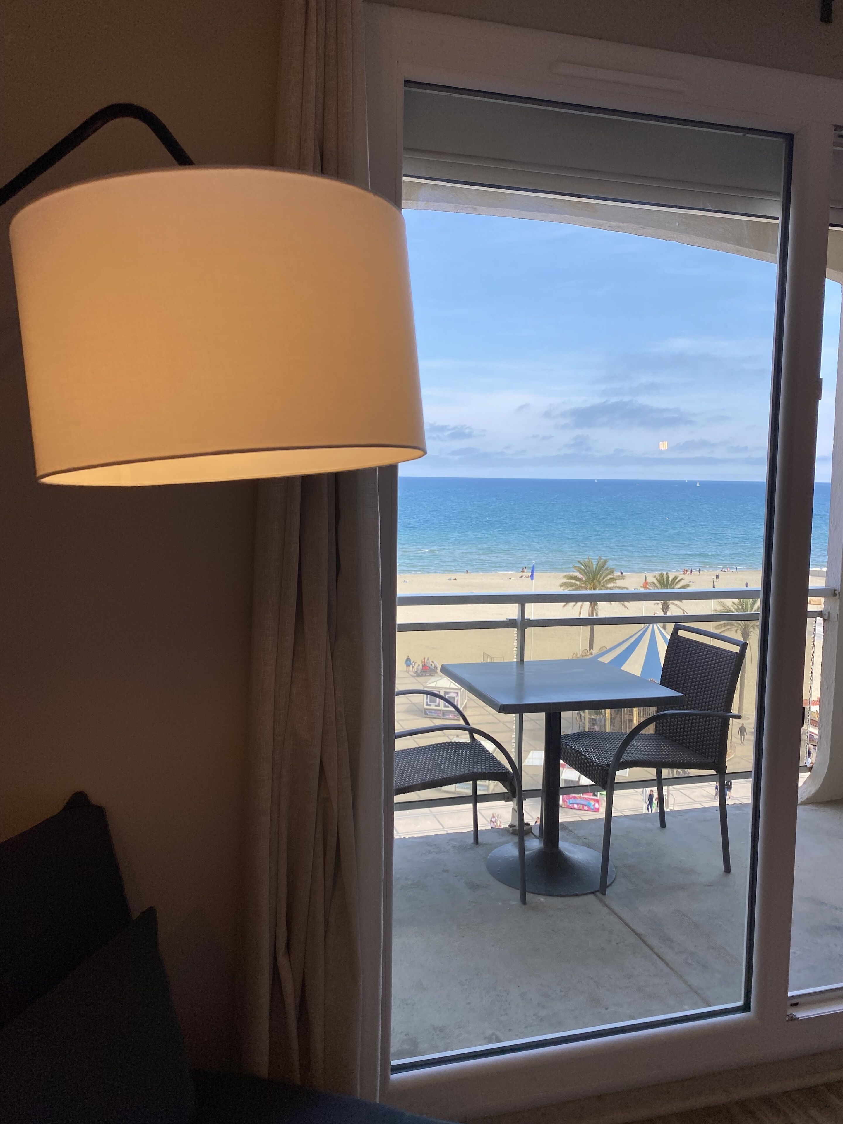 Un balcón con mesa y sillas, con vistas a una playa donde hay gente y un cielo despejado.
