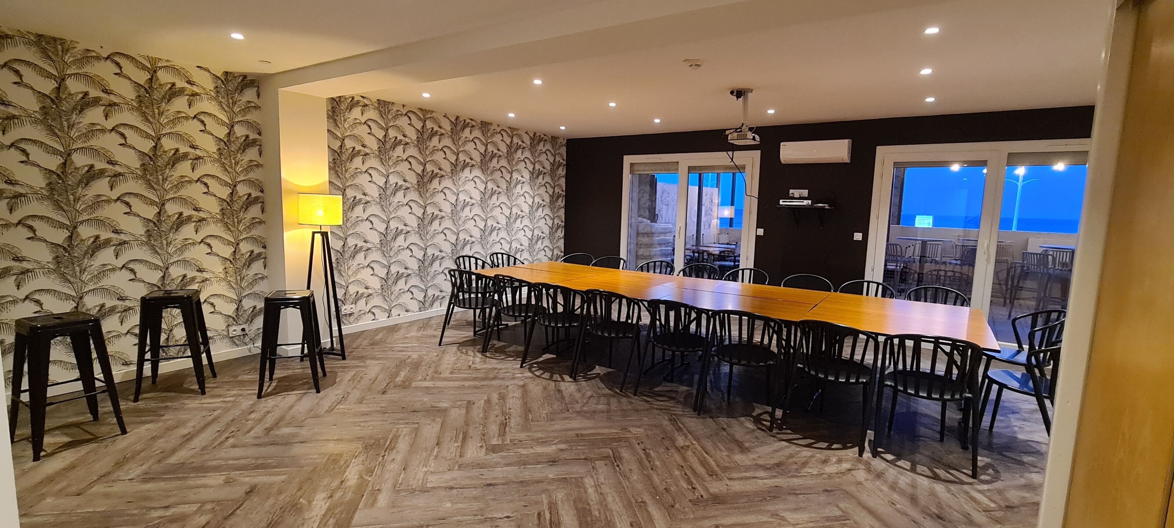 Un coin repas avec une longue table en bois et des chaises noires, un papier peint à motifs et une lampe.