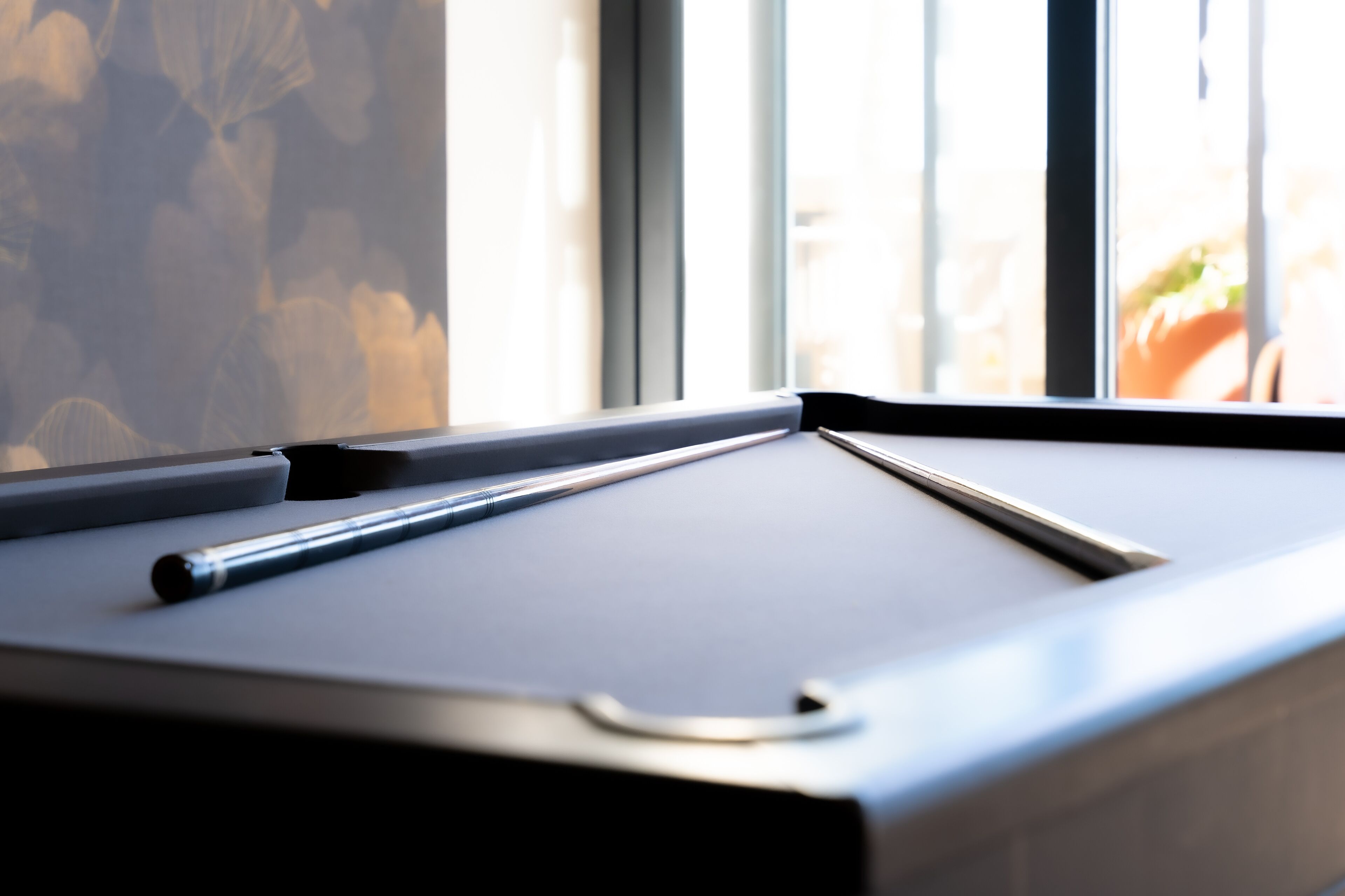 A pool table with a cue stick, near a window with blinds.