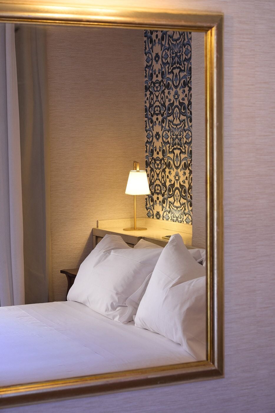 Ett hotellrum med en säng, kuddar, ett nattduksbord, en lampa och en tapet med mönster.