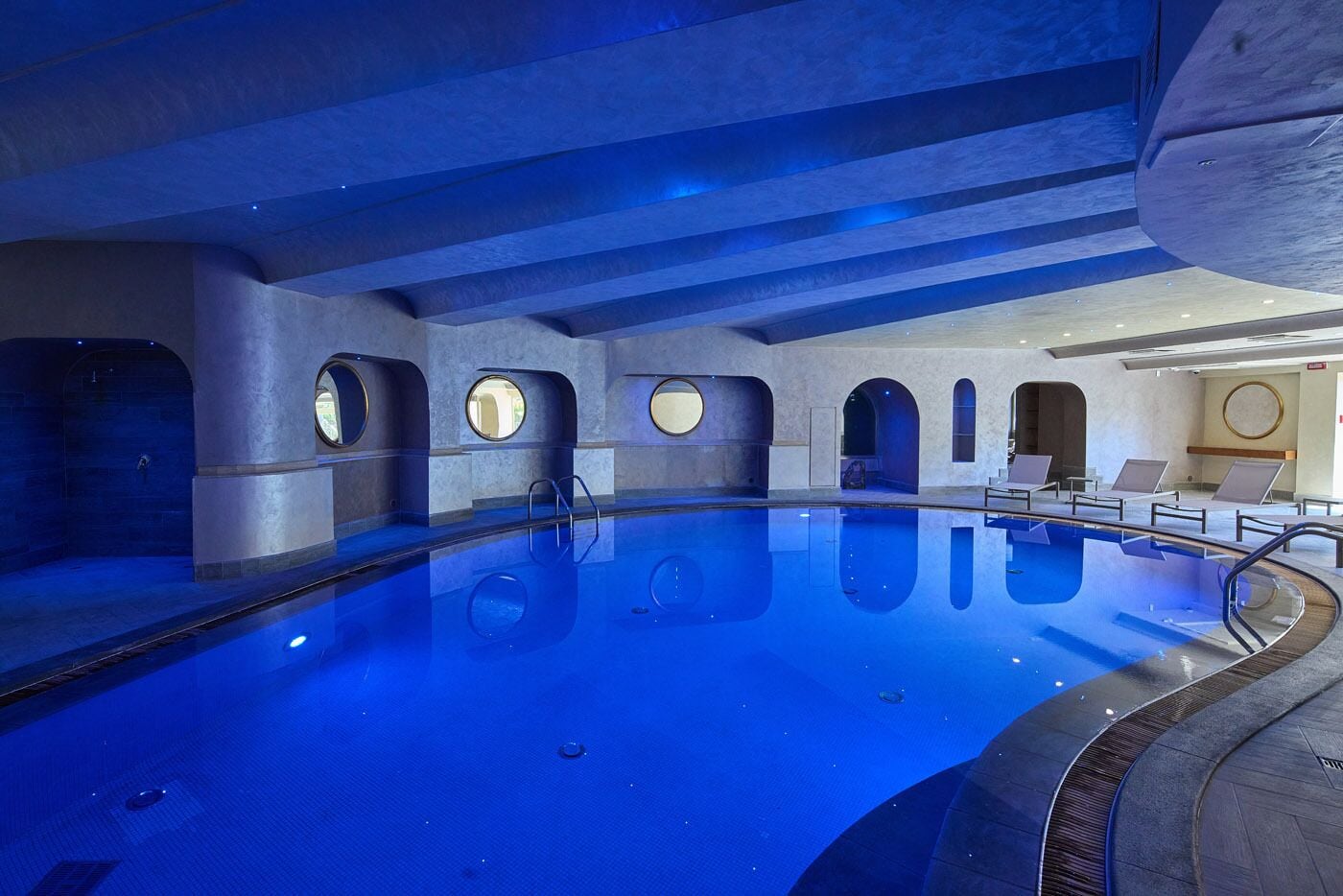 Ein großes, überdachtes Schwimmbecken in Blau, umgeben von rundbogigen Nischen und einer Lounge mit Sesseln.