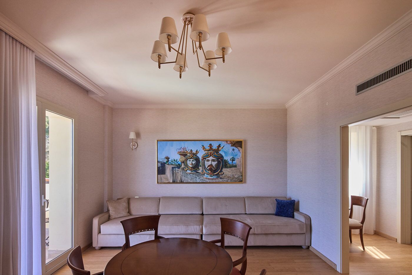 A modern living room with a beige sofa, a dining table with chairs, a chandelier, and a large painting on the wall.