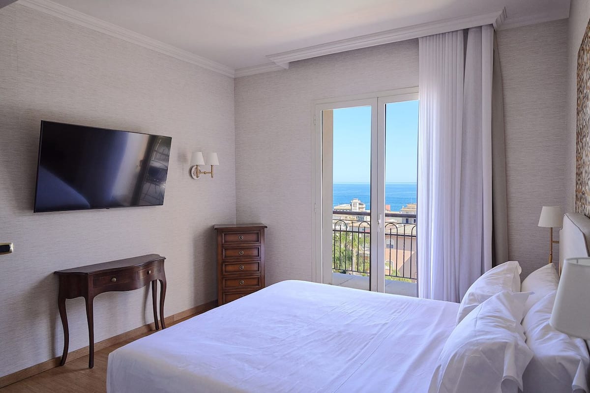A hotel room with a bed, a TV, a dresser, a lamp, and a view of the sea.