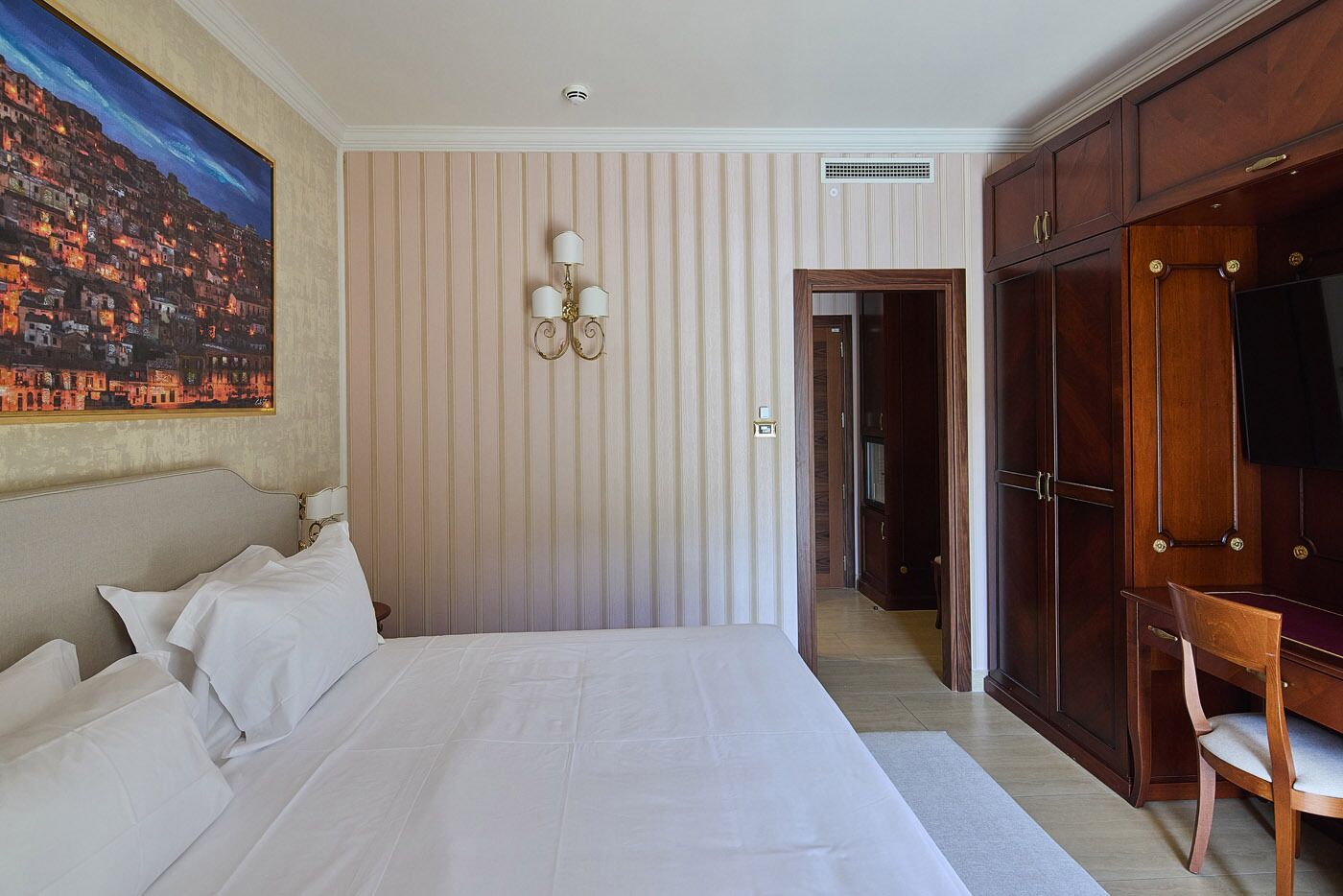 A hotel room with a large bed, a painting, a wardrobe, and a television.