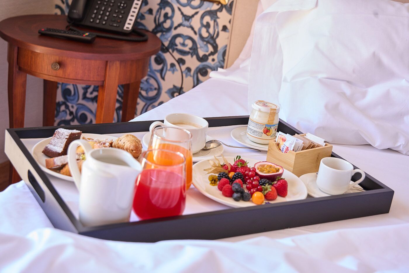 Ein Frühstücksbrett mit Gebäck, Obst, Kaffee und einer Marmeladenglas auf einem Bett.