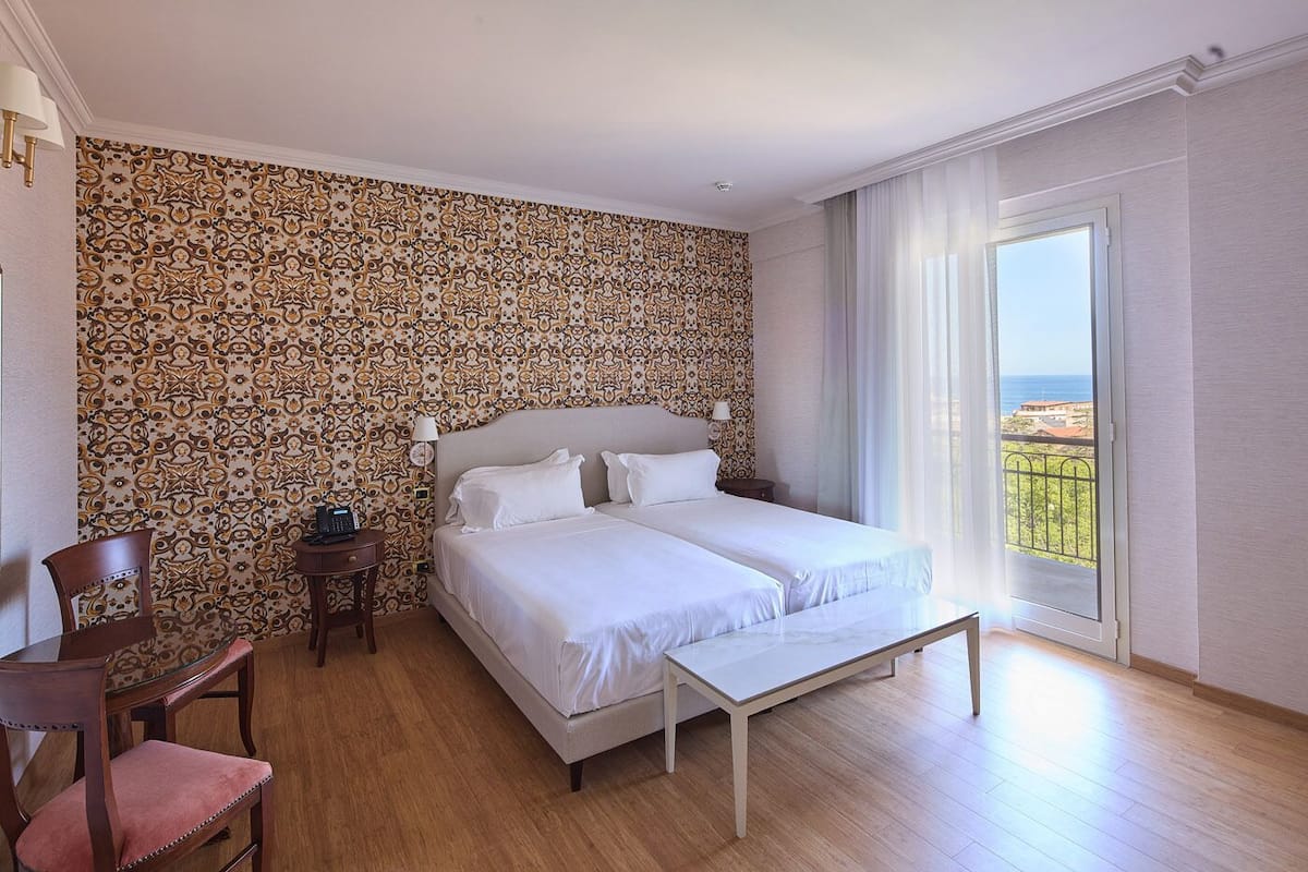 A bedroom with a bed, a desk, a chair, and a view of the sea.