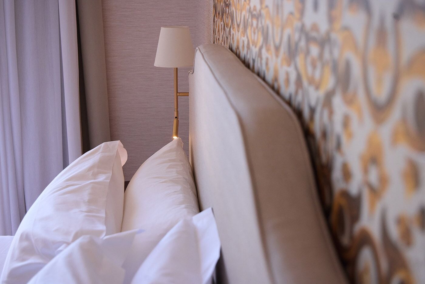 A hotel room with a bed, white pillows, a bedside lamp, and patterned wallpaper.