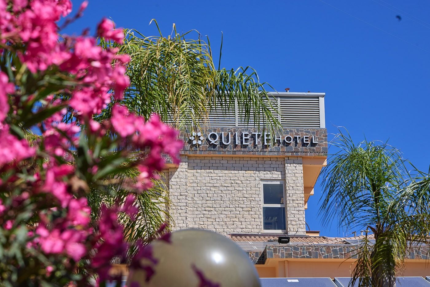Un hôtel à la façade en pierre, une enseigne indiquant « HÔTEL CALME » et un ciel d’un bleu limpide.