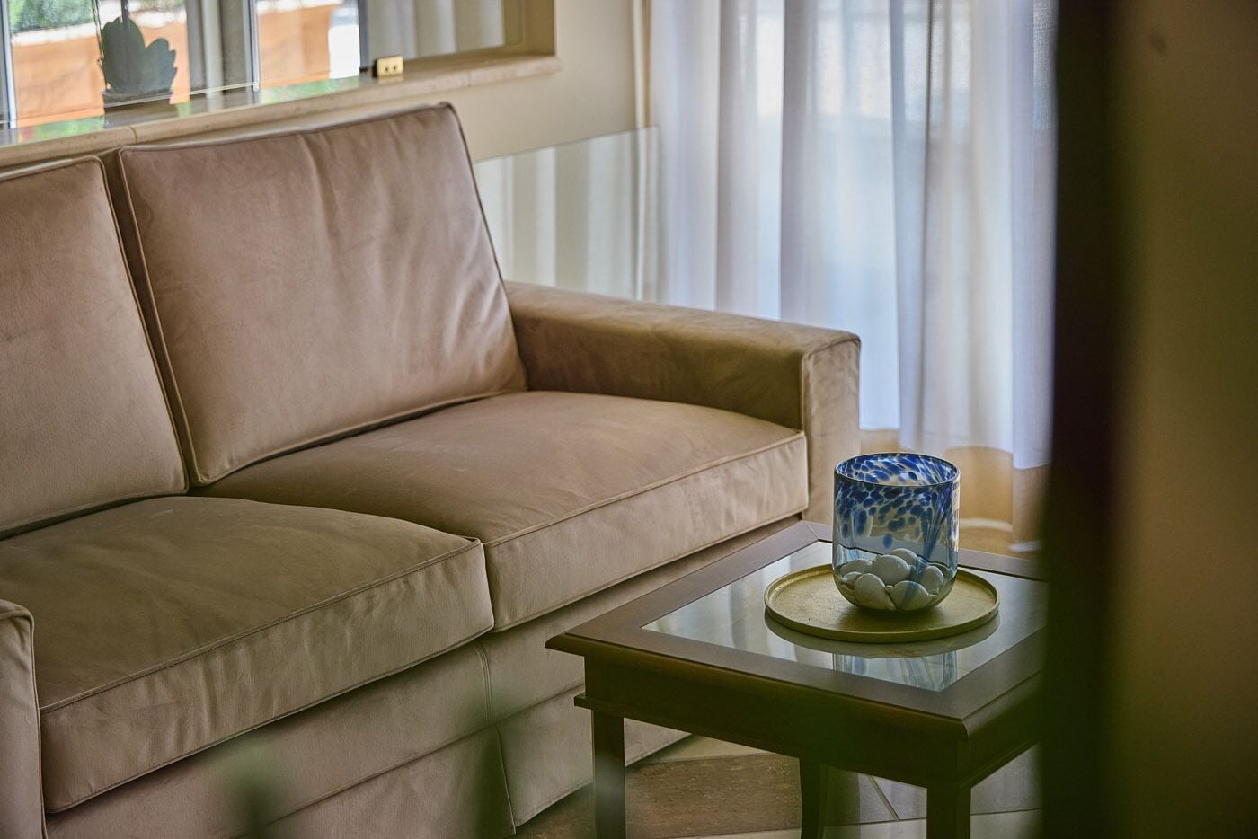 A beige sofa with a glass table beside it, a blue vase with white stones, and a window with sheer curtains.