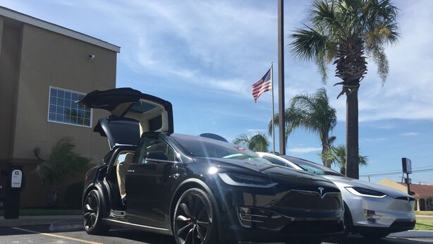 Une Tesla noire est garée sur un parking, son toit ouvrant est relevé. On aperçoit des palmiers et un bâtiment en arrière-plan.