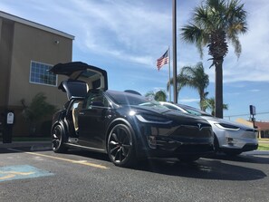Une Tesla noire est garée sur un parking, son toit ouvrant est relevé. On aperçoit des palmiers et un bâtiment en arrière-plan.