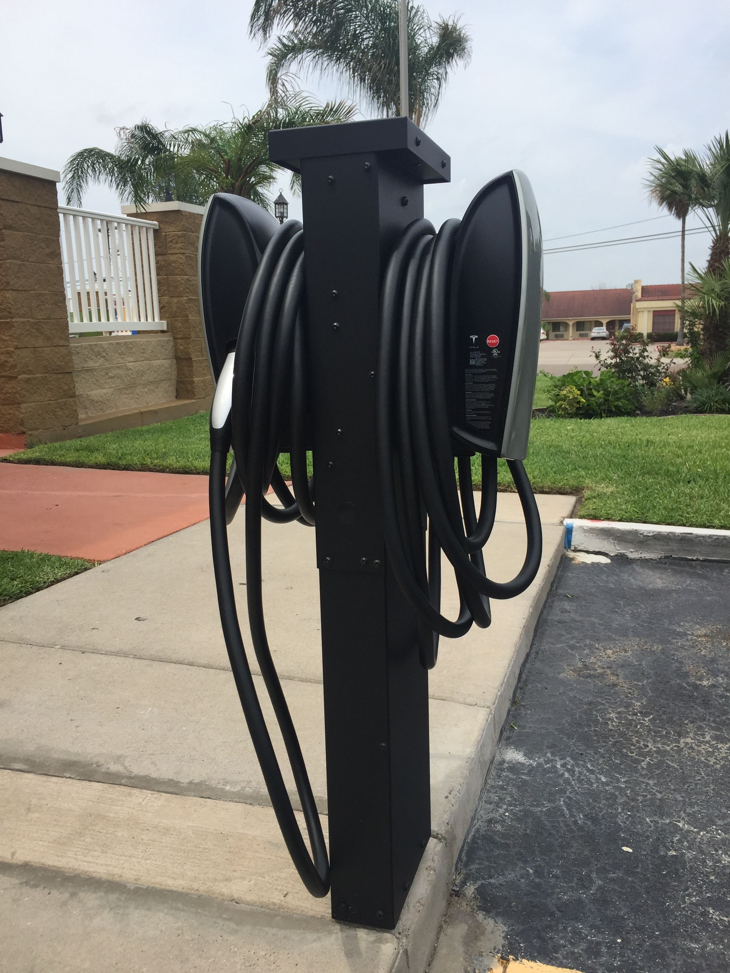 Una estación de carga para vehículos eléctricos negros con varios cables, ubicada en un estacionamiento con un edificio y palmeras al fondo.