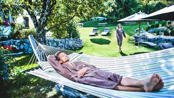 A hammock in a garden with a stone wall, a bench, and a person walking in the background.