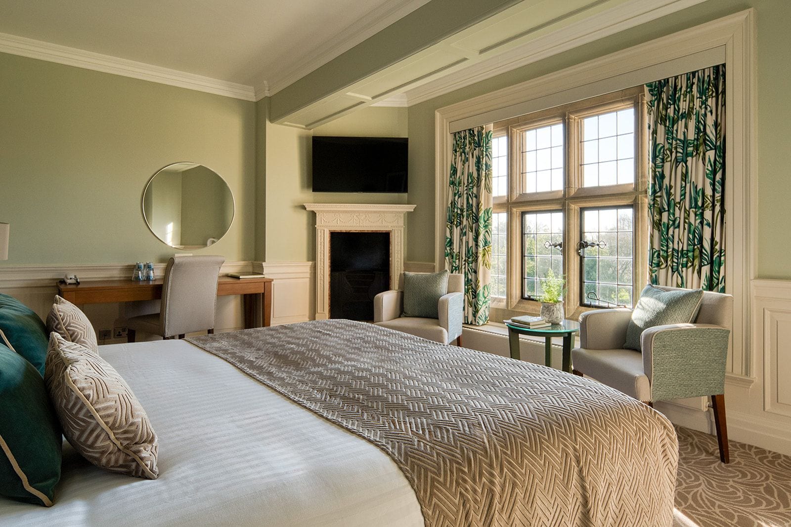 A hotel room with a bed, two armchairs, a desk, a fireplace, and a large window with curtains.