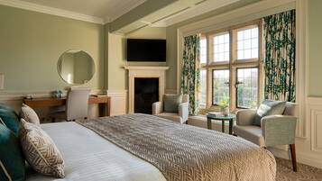 A hotel room with a bed, two armchairs, a desk, a fireplace, and a large window with curtains.