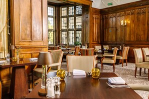 A hotel room with a wooden paneled wall, a large window, and a dining area with chairs and a table set for two.