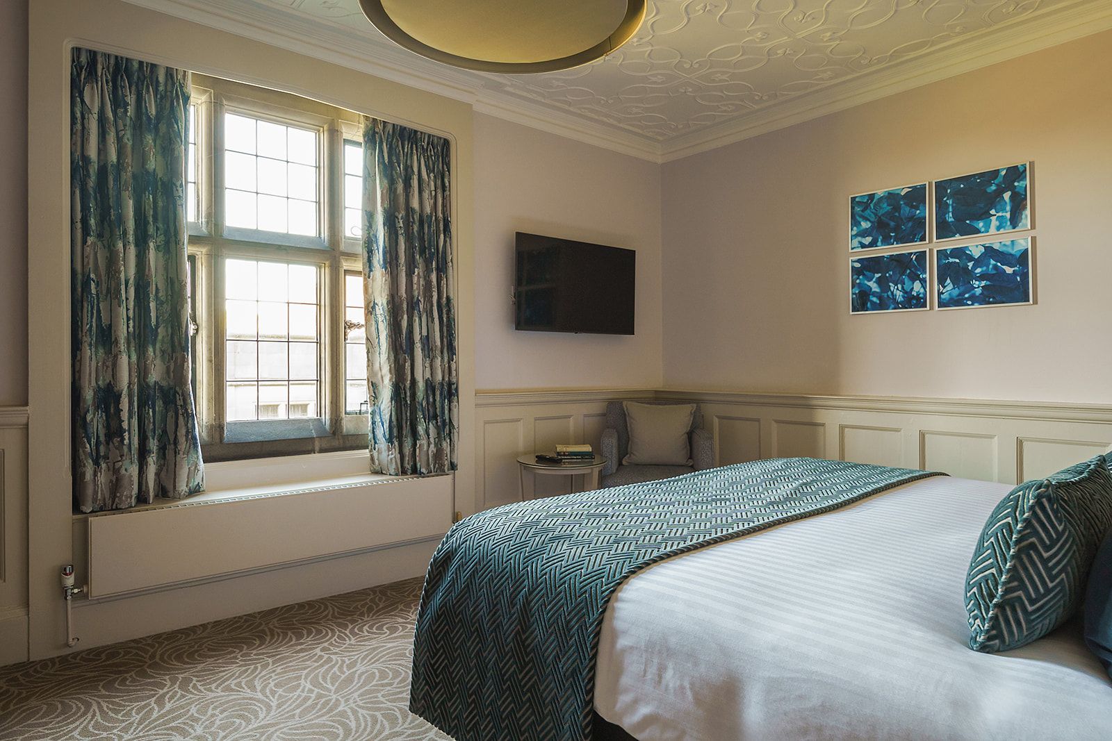 A hotel room with a large bed, a window with curtains, a flat-screen TV, and artwork on the wall.