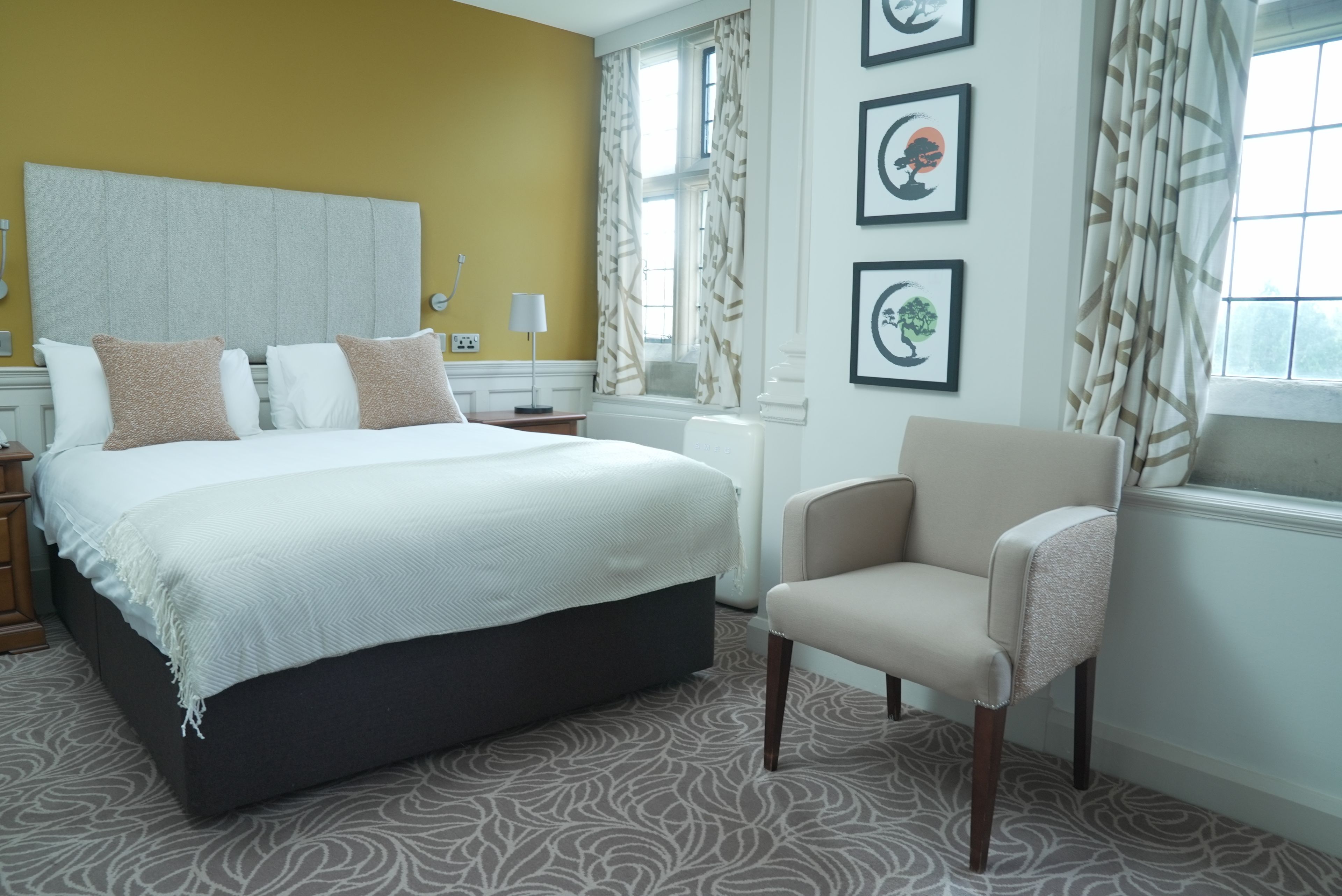 A hotel room with a bed, a chair, a window, and artwork on the wall.