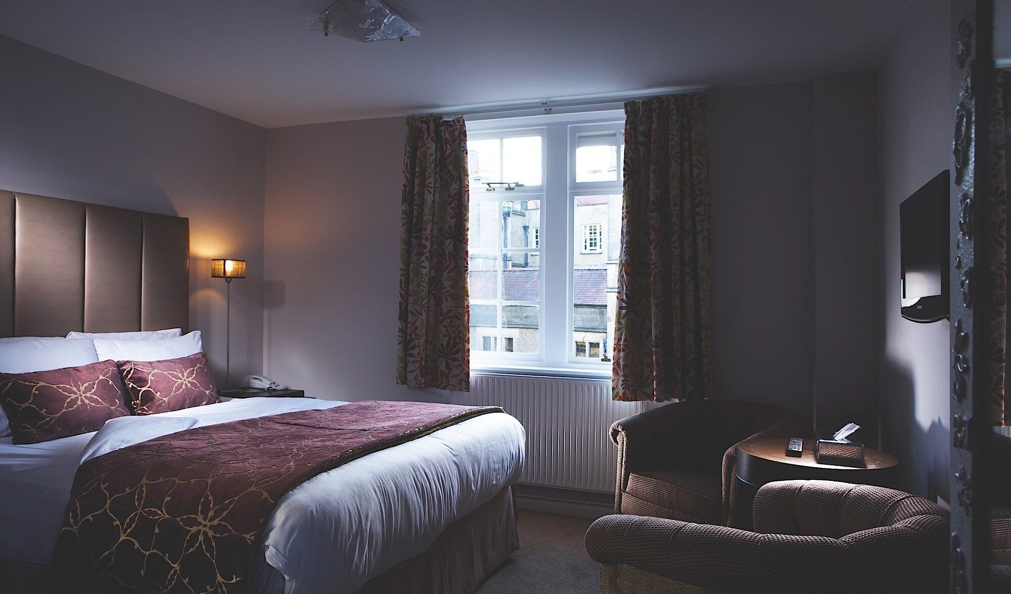 Una habitación de hotel con cama, una silla, una mesita, una lámpara y una ventana con cortinas.