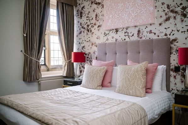 A bedroom with a large bed, two bedside tables, a window with curtains, and a floral wallpaper.