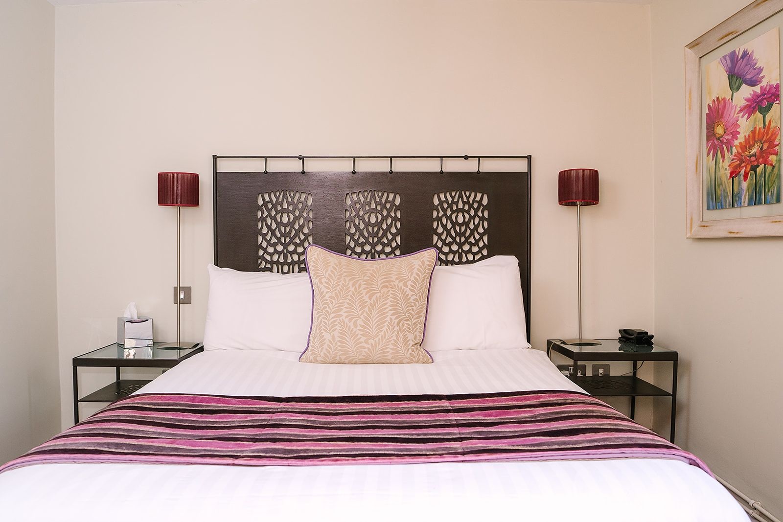 Una habitación de hotel con cama, dos mesitas de noche, lámpara, un cuadro y un cabecero decorativo.