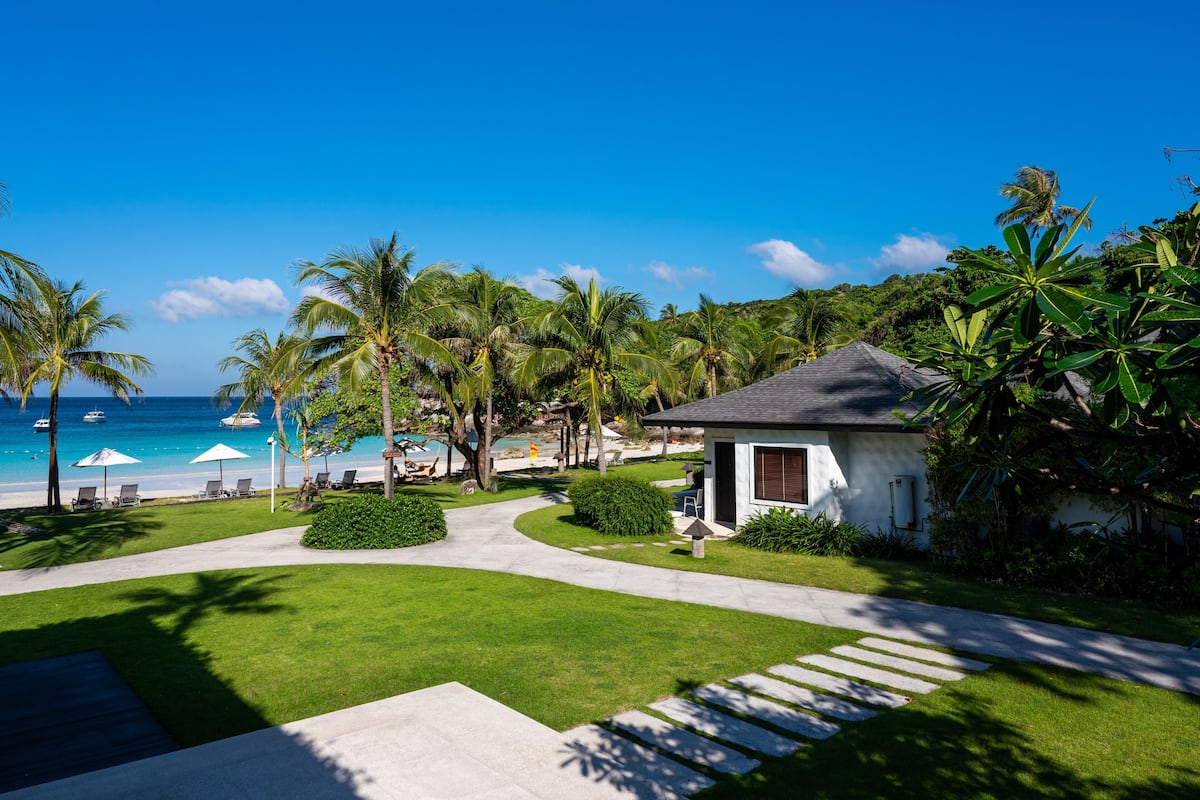 A resort with a clear view of the ocean, palm trees, and a pathway leading to a building.