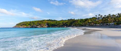 A beach with clear turquoise water, a sandy shore, and a hill with greenery and buildings in the background.