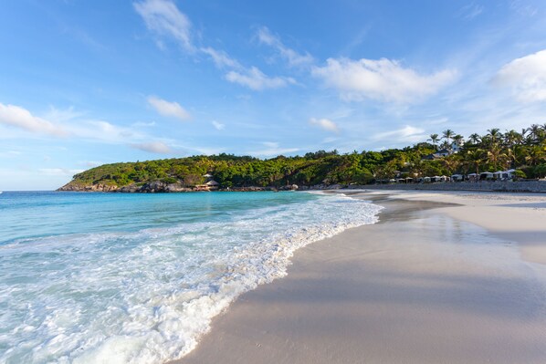 A beach with clear turquoise water, a sandy shore, and a hill with greenery and buildings in the background.