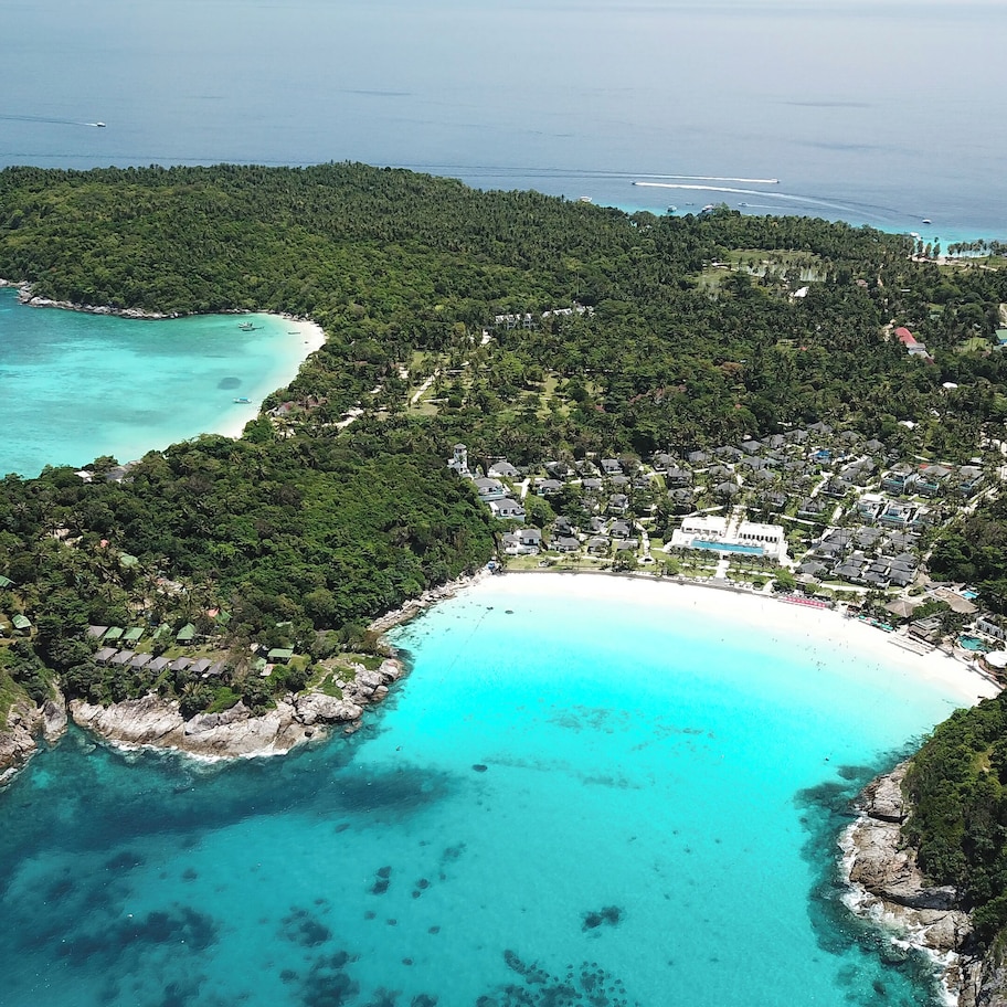 An aerial view of a coastal resort with a central building surrounded by lush greenery and turquoise waters.