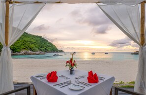 A table set for two with a view of a beach and boats at sunset.